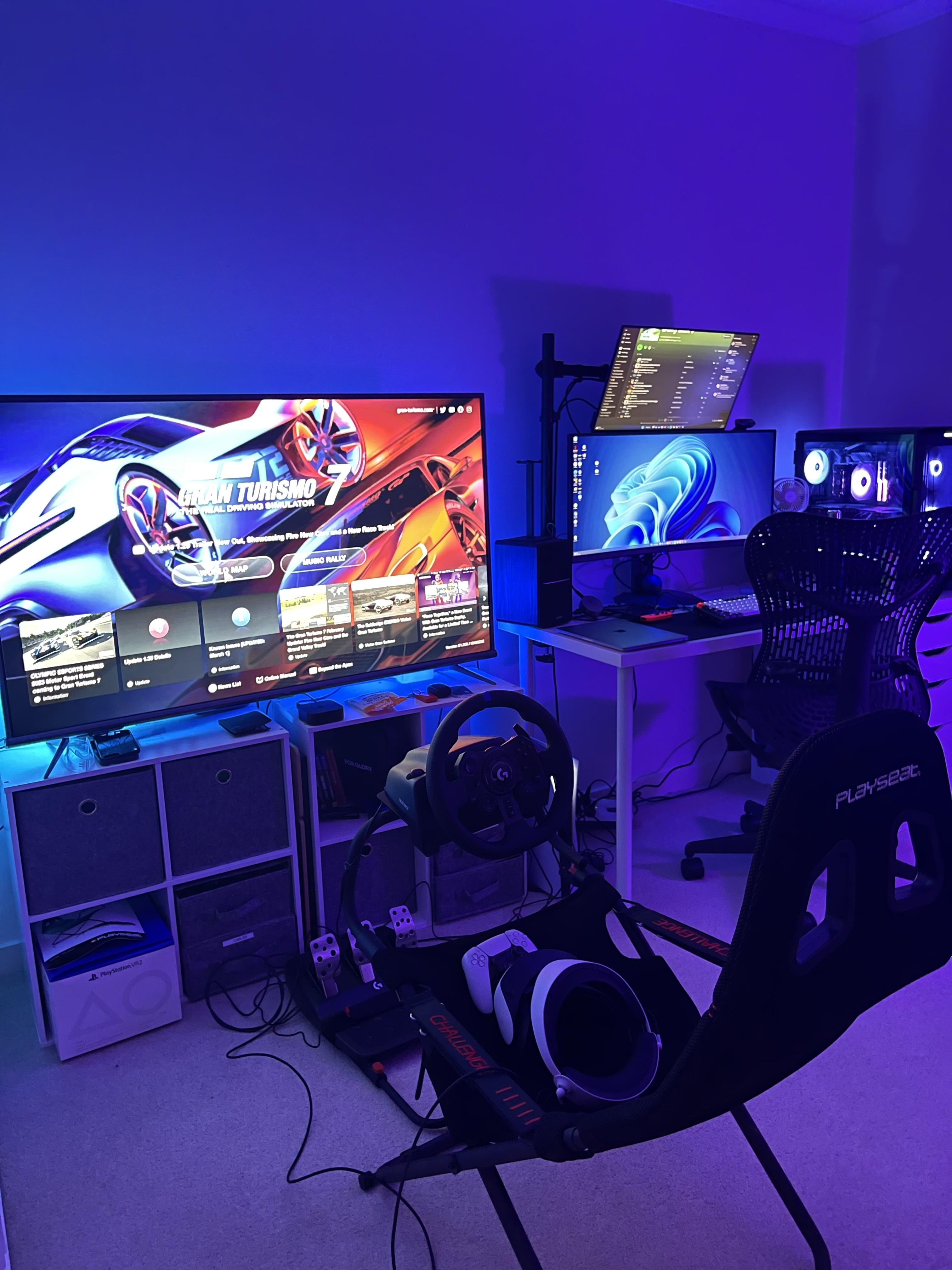 A racing simulator rig with a wheel, pedals, and seat facing a TV, alongside a PS5, a VR headset, and a separate multi-monitor desk