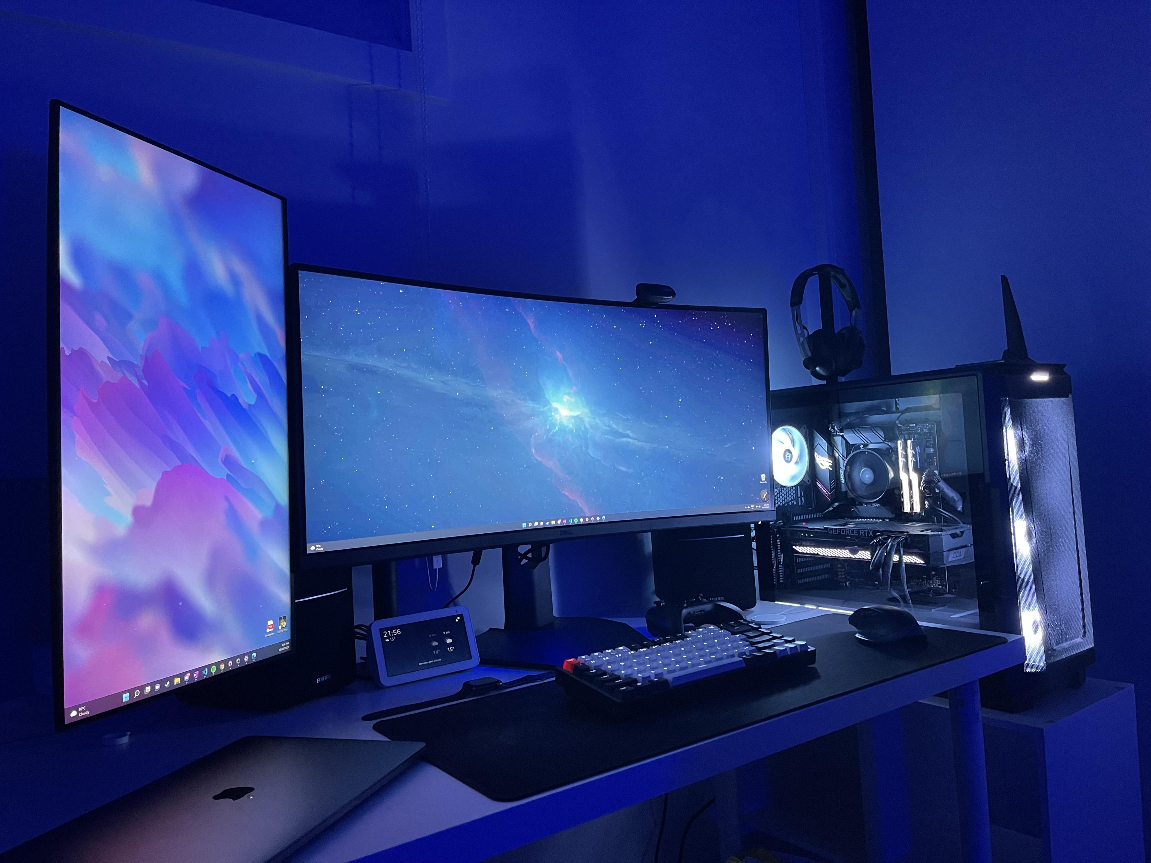 A dual-monitor desk setup under blue ambient lighting, with a mechanical keyboard and an open PC case showing RGB-lit components